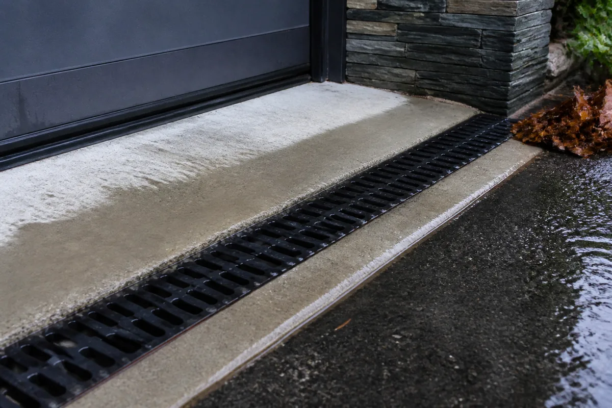 Caniveau à grille scellé en pied de descente devant une porte de garage, béton clair humide de pluie