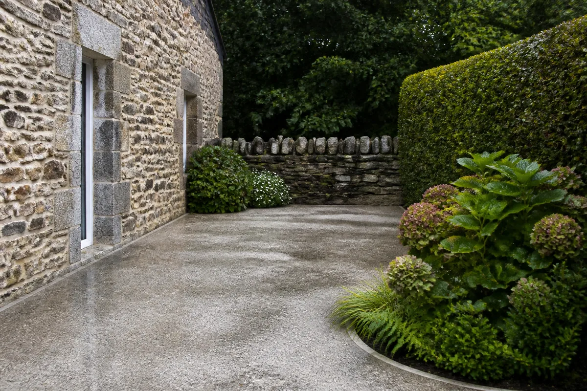 Dalle de cour en béton désactivé gris clair bordée d’un muret de schiste devant une longère du sud Finistère