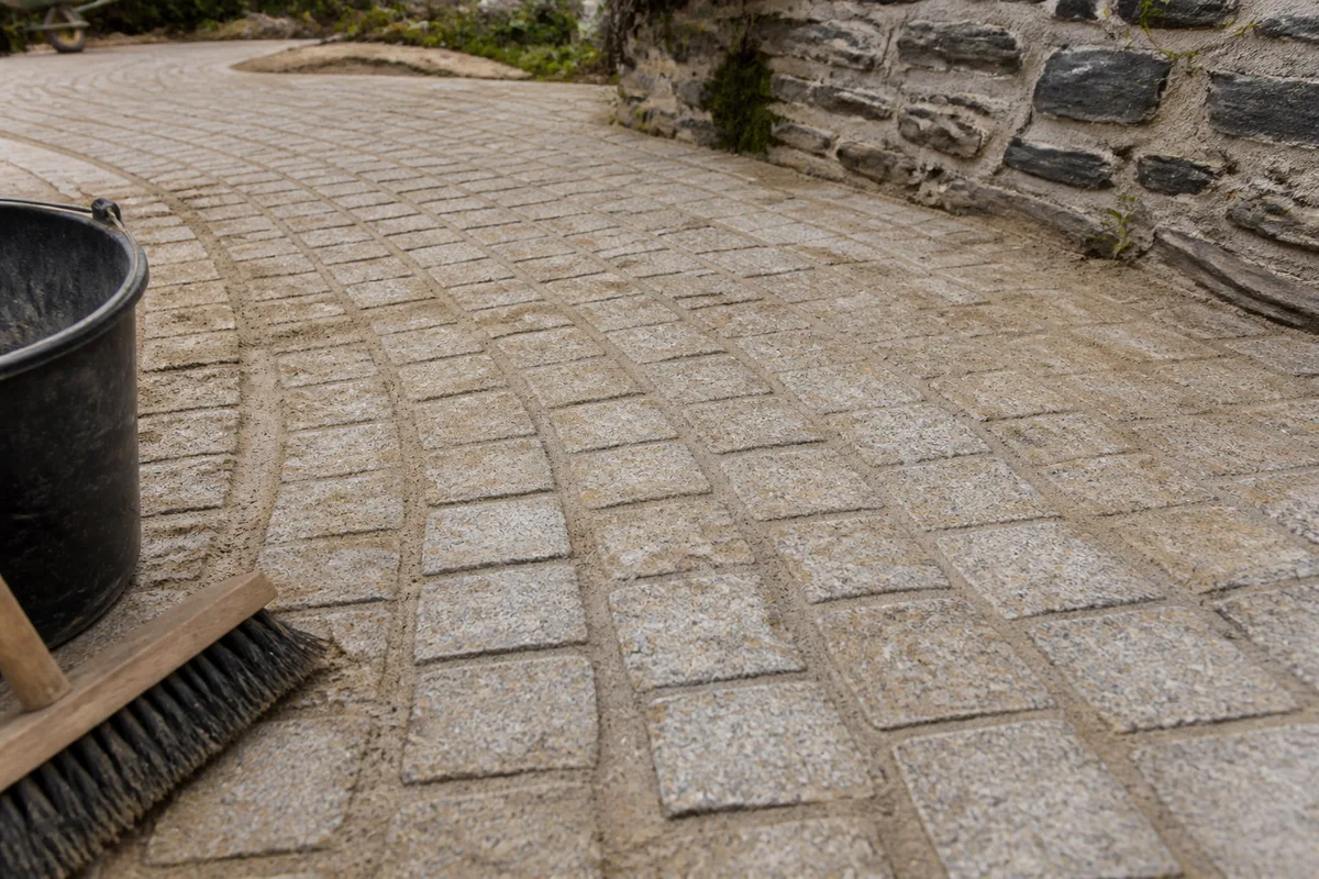 Pavé de granit clair posé en rangs légèrement courbes devant une maison de pierre, joints de sable encore frais